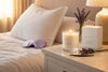 Luxury lavender vanilla scented candle by Safispa in a glass jar on a white nightstand, glowing softly beside an open book, sleep mask, and dried lavender in a cozy bedroom setting.