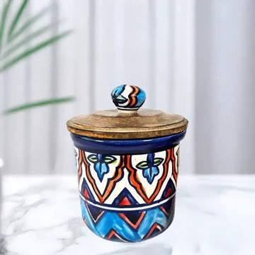 Ceramic jar with colorful geometric design and wooden lid on a white surface with a plant in the background