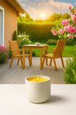 Candle in a white holder on a white background with a garden scene featuring a wooden table and chairs in the background.