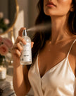 Woman applying SaFiSpa Belle Rose Body Mist to her neck during a luxury self-care routine, featuring the authentic Belle Rose body spray bottle with a soft floral fragrance, warm natural light, and elegant beauty styling.