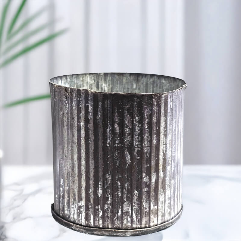 Silver ribbed metal planter on a white surface with a blurred green plant in the background