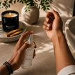 Adult applying SaFiSpa Palo Santo Body Mist to the wrist in a luxury spa-inspired setting with a lit candle, showcasing a unisex woody body spray for everyday self-care, refreshing fragrance, and clean modern grooming.