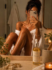 SaFiSpa Hibiscus Body Oil mirror selfie lifestyle image featuring glowing hydrated skin, luxury body care routine, non-greasy body oil, radiant-looking legs and shoulders, warm bathroom lighting, and premium self-care aesthetic.