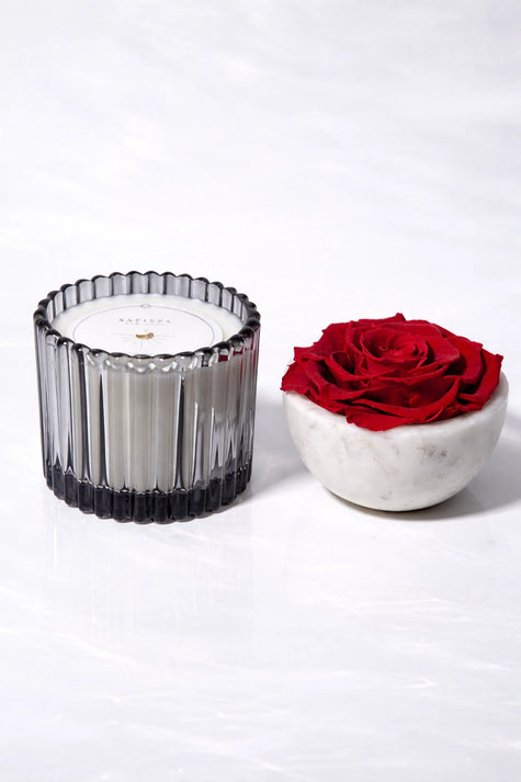 Candle in a ribbed glass holder next to a red rose in a white marble bowl on a white background