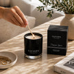 A black glass candle jar with the brand name SAFISPA New York is lit with a wooden match by a person's hand, next to its black packaging box labeled 'Safispa New York Passion Spice'. 